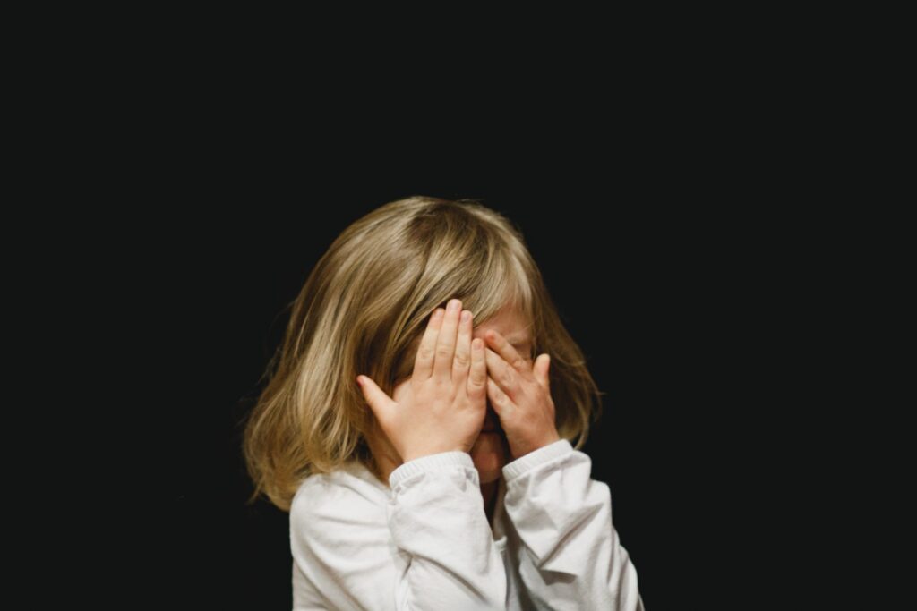 Young girl with her hands covering her face as if to hide standing against a black background.